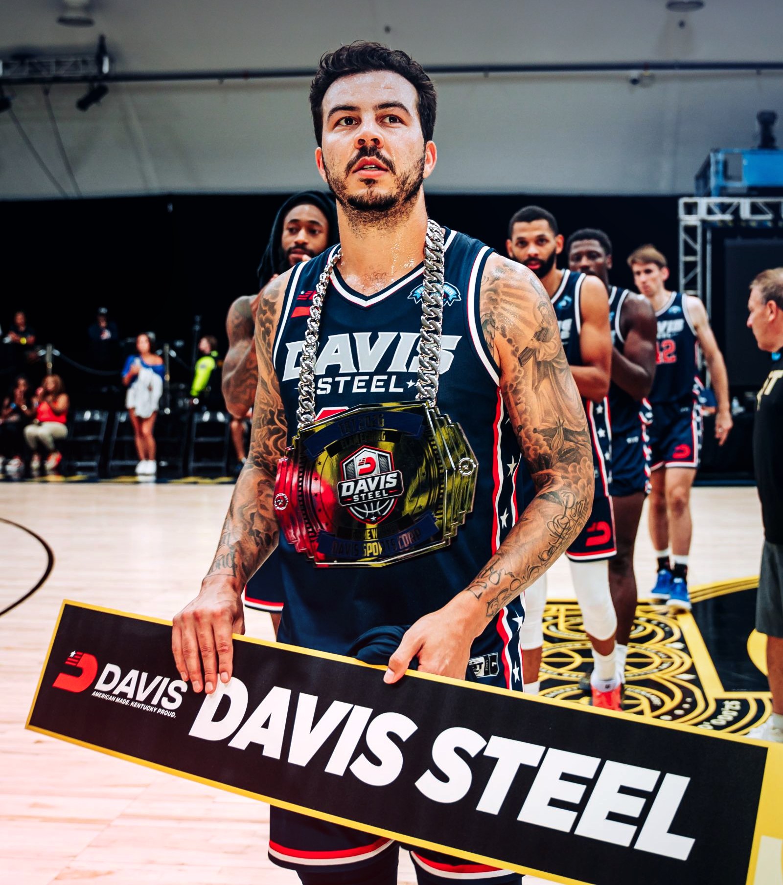 A Davis Steel player wearing the Elam Ending chain and holding the Davis Steel sign, with teammates lined up behind him.